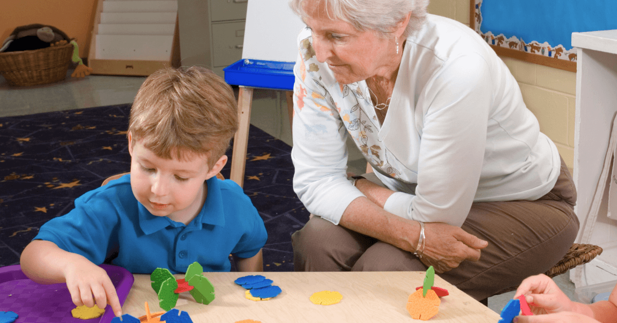An older woman uses observation techniques as she watches a young boy in a blue shirt play with colorful foam shapes on a table in a classroom setting. Another child's arm is partially visible beside them.