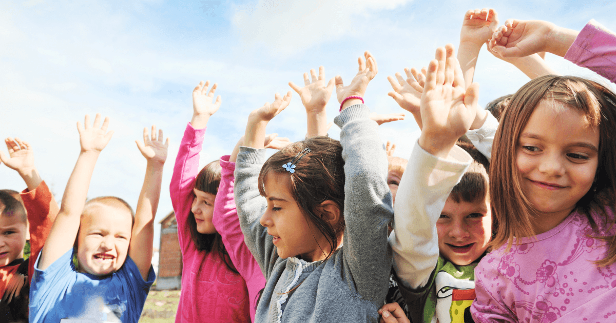 A group of young children stand outdoors with their arms raised, smiling and appearing joyful under a blue sky, showcasing the fun and camaraderie found in many preschool options.