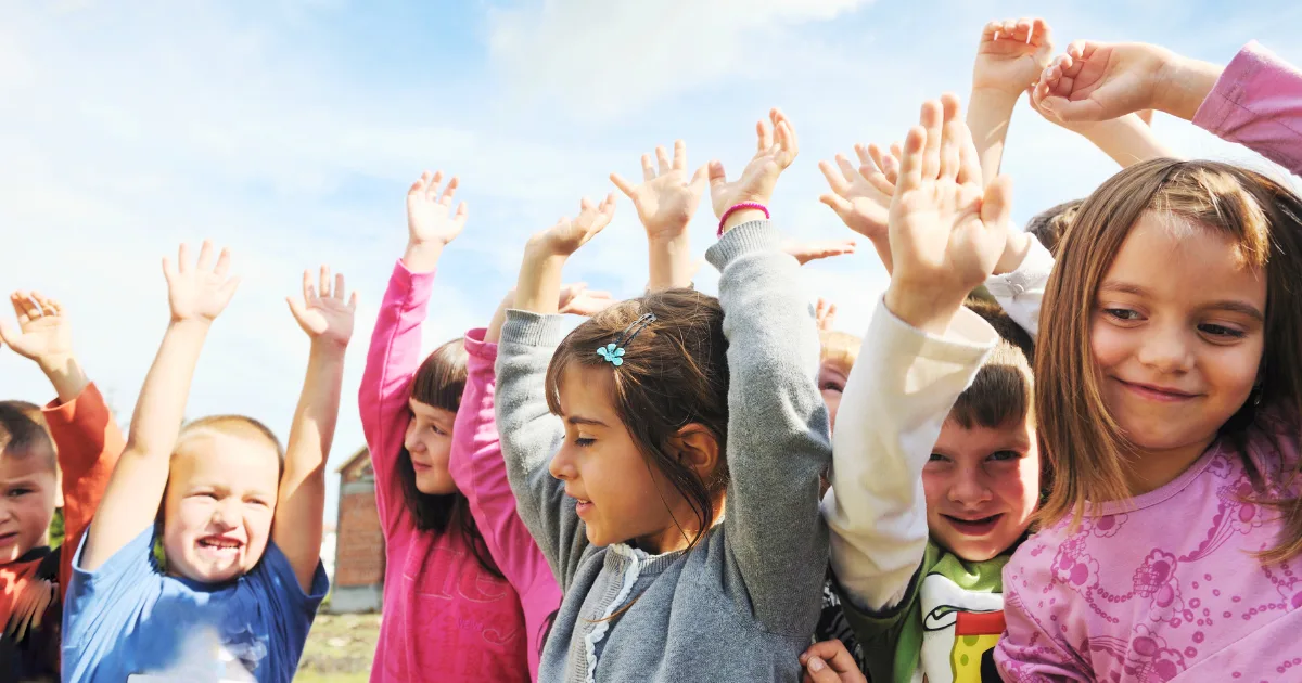 A group of young children stand outdoors with their arms raised, smiling and appearing joyful under a blue sky, showcasing the fun and camaraderie found in many preschool options.