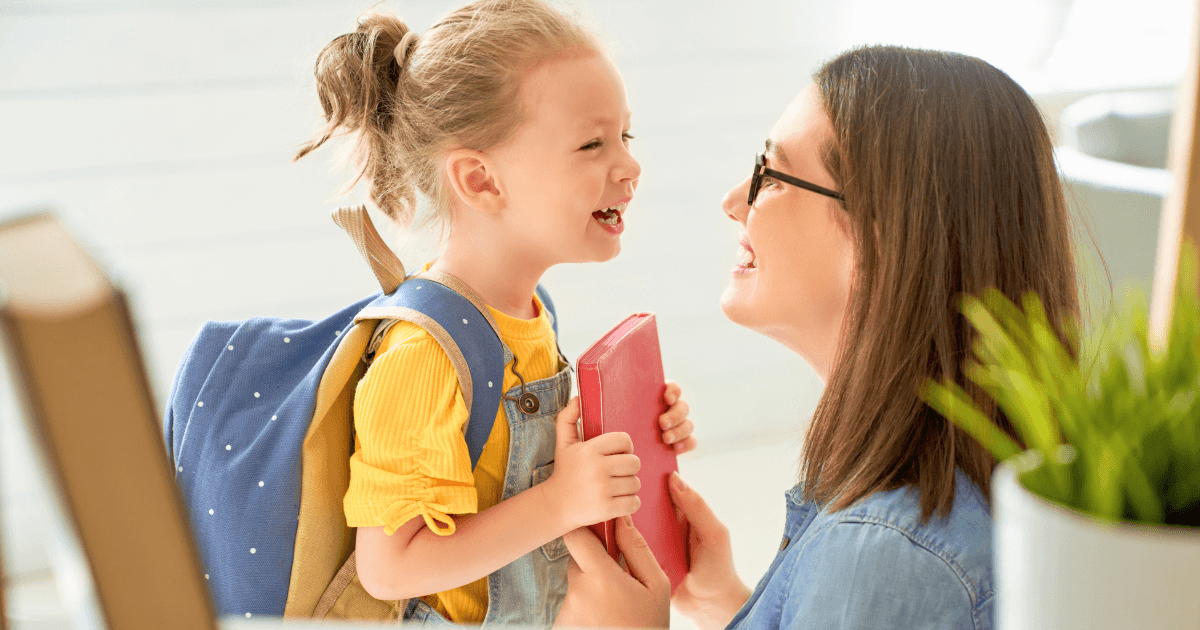 A smiling child with a backpack is holding a red book and facing an adult woman in glasses. They are laughing together indoors, with sunlight streaming in as they look over a parent handbook.