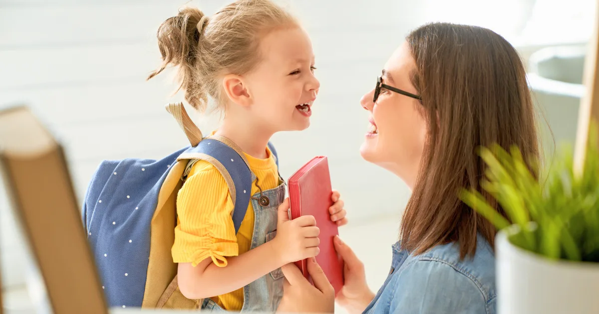 A smiling child with a backpack is holding a red book and facing an adult woman in glasses. They are laughing together indoors, with sunlight streaming in as they look over a parent handbook.