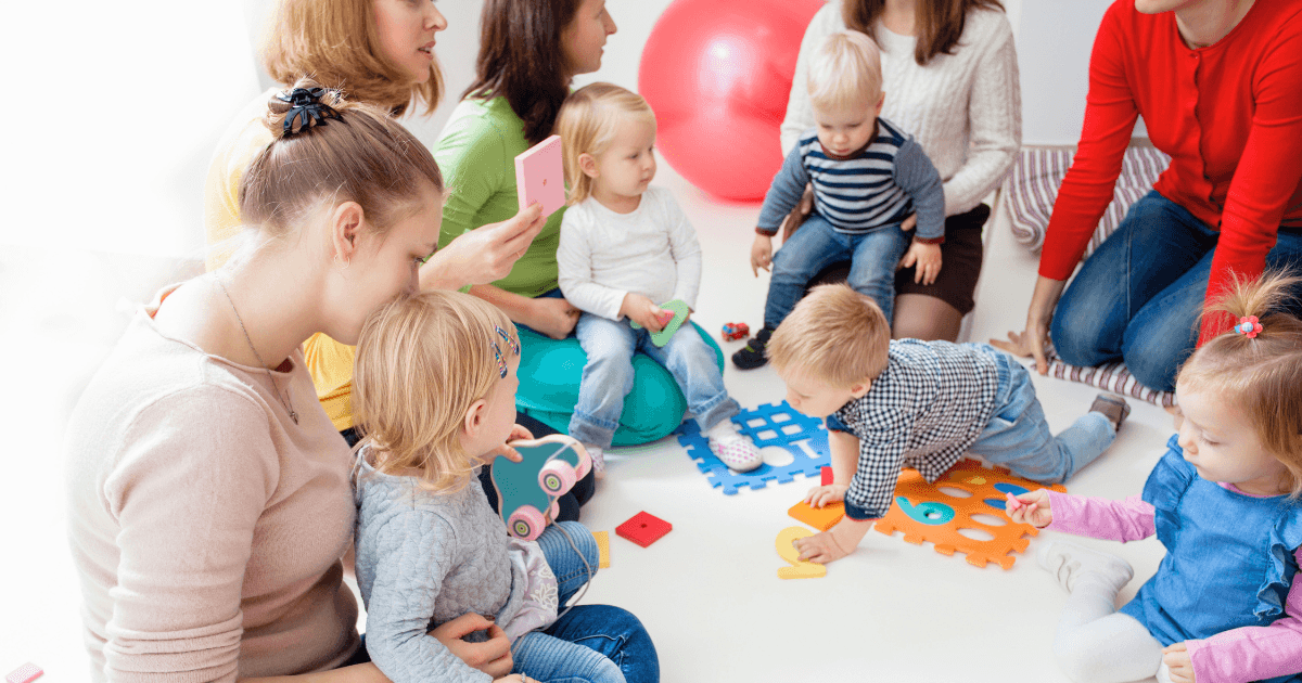 A group of young children and adults, part of parent volunteer programs, sit and play together on the floor with colorful foam puzzle pieces and toys in a bright, cheerful room.