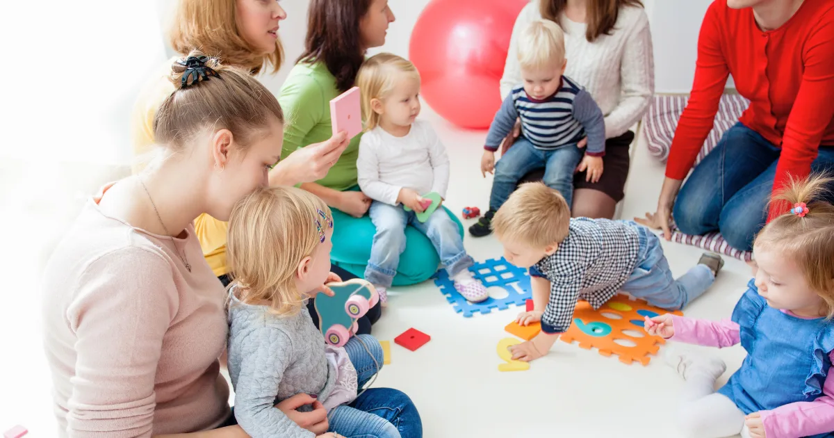 A group of young children and adults, part of parent volunteer programs, sit and play together on the floor with colorful foam puzzle pieces and toys in a bright, cheerful room.