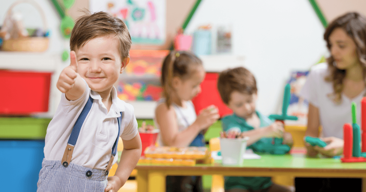 A smiling young boy gives a thumbs-up in a colorful classroom, showcasing the success of program marketing as other children and a teacher work on crafts at a table in the background.