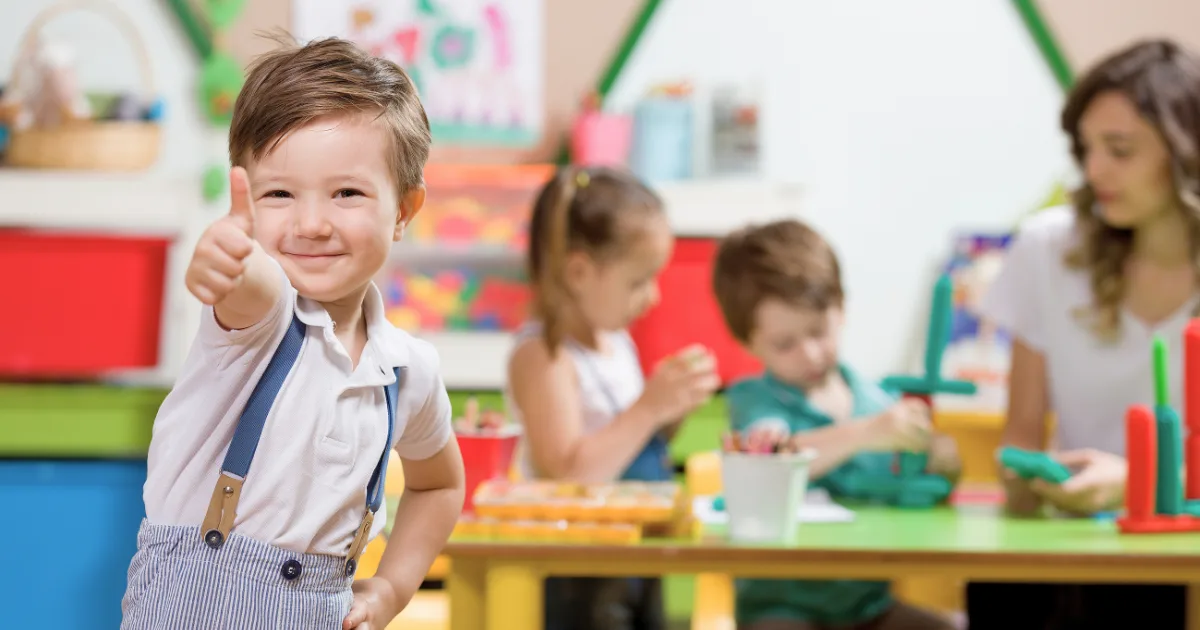 A smiling young boy gives a thumbs-up in a colorful classroom, showcasing the success of program marketing as other children and a teacher work on crafts at a table in the background.