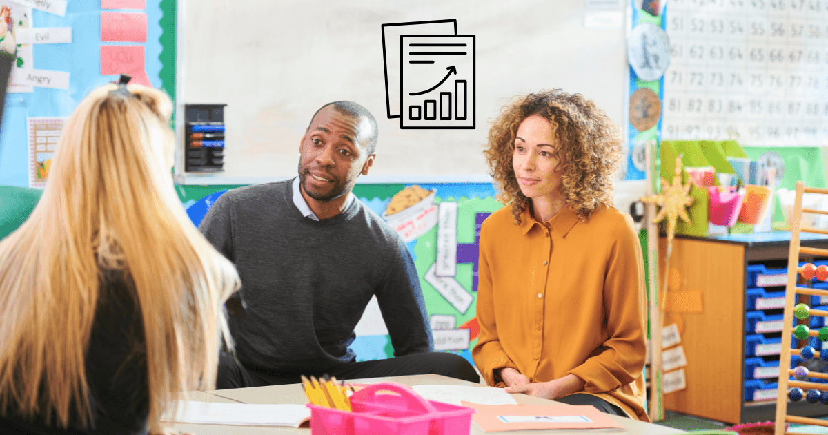 Two adults sit across from a student in a colorful classroom, engaged in conversation. Charts and progress reports decorate the background, suggesting a parent-teacher meeting or academic discussion.