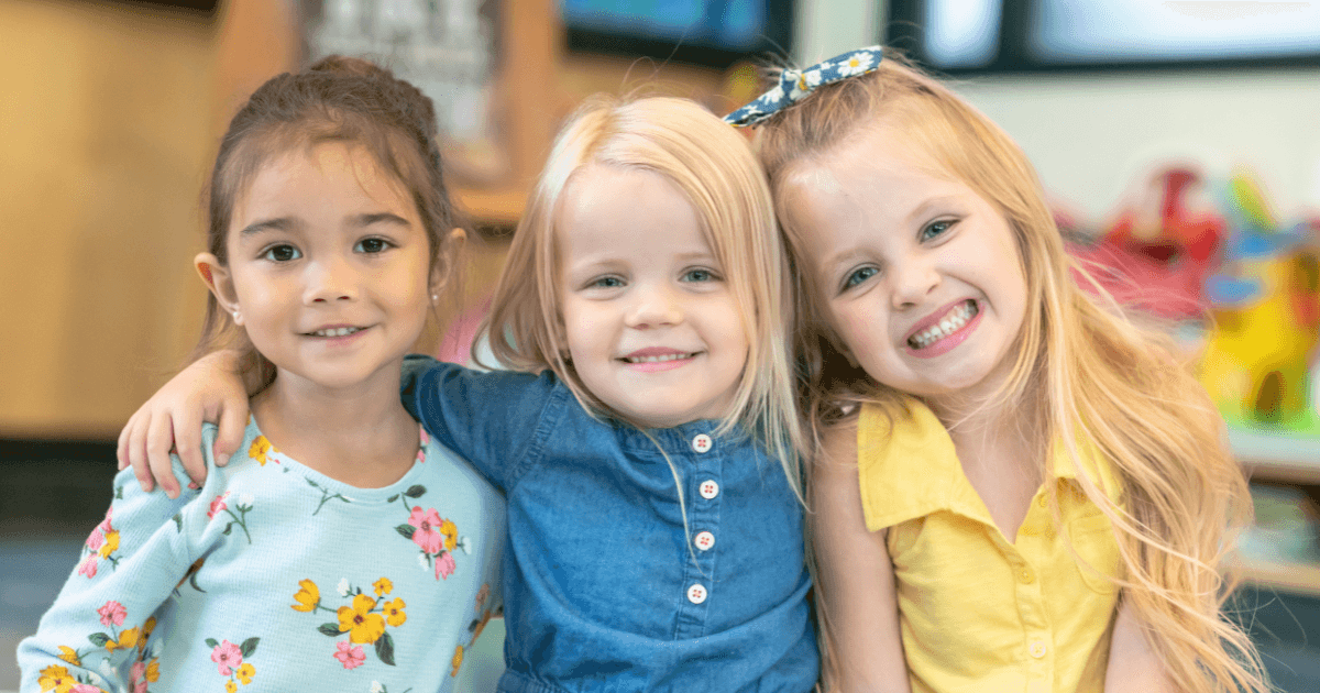 Three young girls are smiling and hugging each other, each in a colorful outfit. The softly blurred classroom setting highlights key preschool readiness signs: social bonding, joy, and comfort in a group environment.