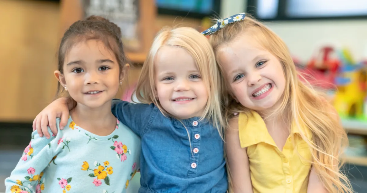 Three young girls are smiling and hugging each other, each in a colorful outfit. The softly blurred classroom setting highlights key preschool readiness signs: social bonding, joy, and comfort in a group environment.