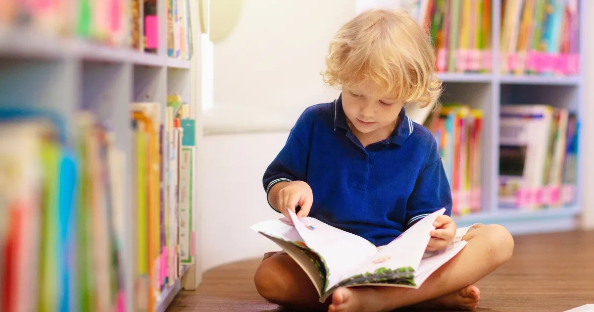 A young child with blonde hair, wearing a blue shirt, sits cross-legged on the floor in a library, reading a colorful picture book about seasonal books, surrounded by shelves filled with books.
