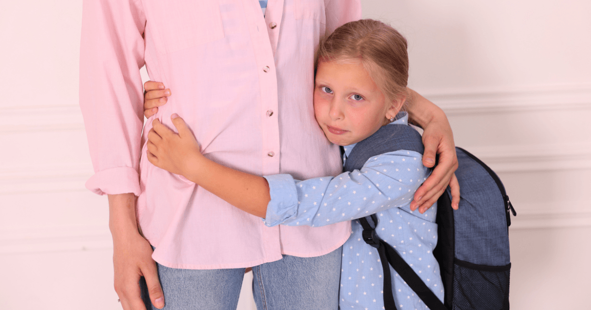 A young girl with a backpack, experiencing preschool separation anxiety, hugs an adult, looking sad or anxious. The adult stands with one hand by their side and the other gently holding the girl. Both are indoors against a light-colored wall.