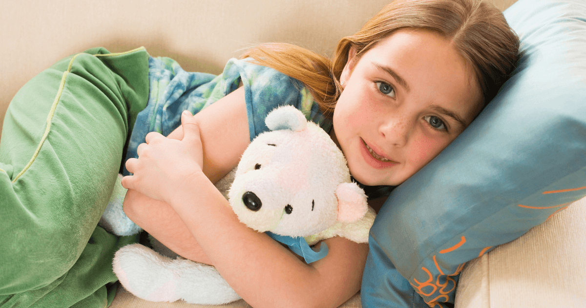A young girl lies on a couch, hugging a white stuffed bear—her cherished transitional object—and resting her head on a blue pillow. She has light brown hair and wears a green and blue shirt with green pants, looking content and relaxed.