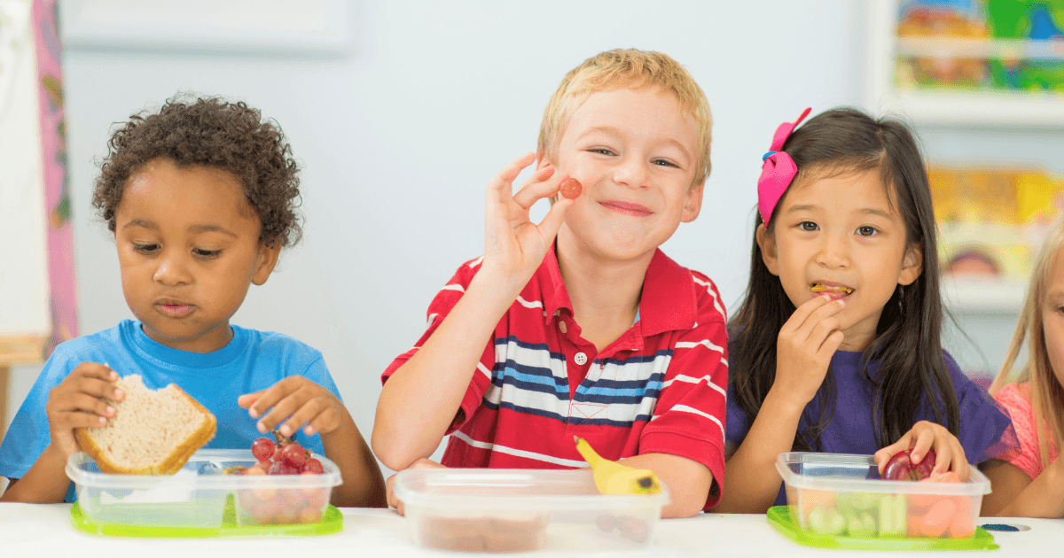 Three young children sit at a table eating lunch from plastic containers, enjoying fresh food together. The boy in the middle smiles while holding a grape, embracing wellness, as his friends also enjoy their healthy meal.
