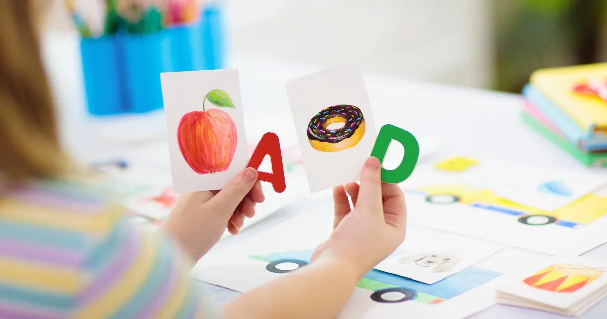 A child holds two cards—one showing an apple with the letter A, and another featuring a donut and the letter D. More preschool worksheets and colorful objects are scattered on the table in the background.