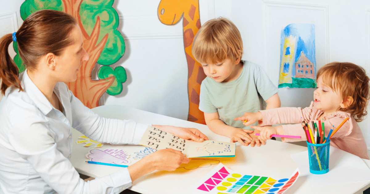 A woman shows a book to two young children at a table with art supplies, colorful drawings, and paper cutouts around them in a bright, playful classroom setting—helping prevent the summer slide through engaging activities.