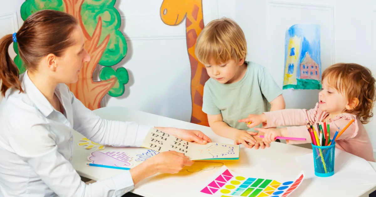 A woman shows a book to two young children at a table with art supplies, colorful drawings, and paper cutouts around them in a bright, playful classroom setting—helping prevent the summer slide through engaging activities.
