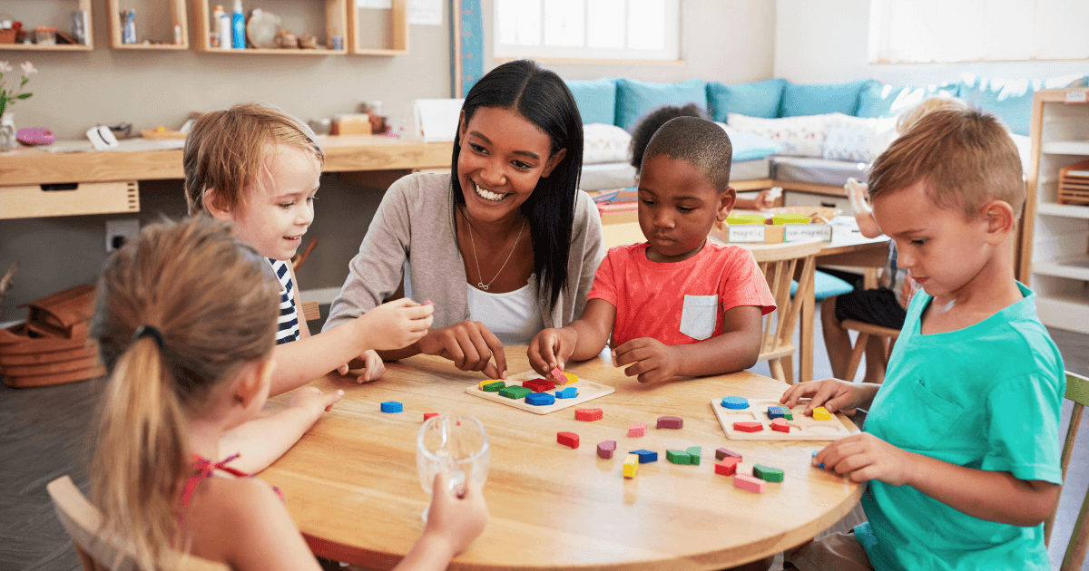 A teacher, engaged in professional development, sits at a round table with four young children, smiling as they play with colorful geometric blocks and shapes in a bright classroom setting.