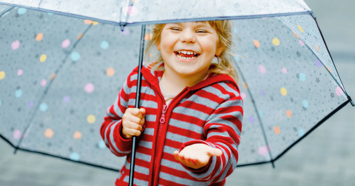 A smiling child in a red striped jacket holds a transparent umbrella with colorful dots, reaching out to catch raindrops—perfect inspiration for rainy day preschool activities.