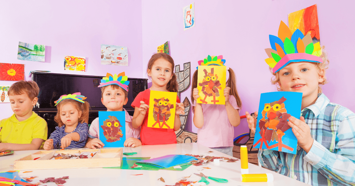 Six young children sit at a table in a colorful classroom, wearing handmade feather headbands and proudly holding up seasonal preschool crafts—owl creations made from paper and leaves. Art supplies and scattered leaves fill the table, while artwork decorates the walls.