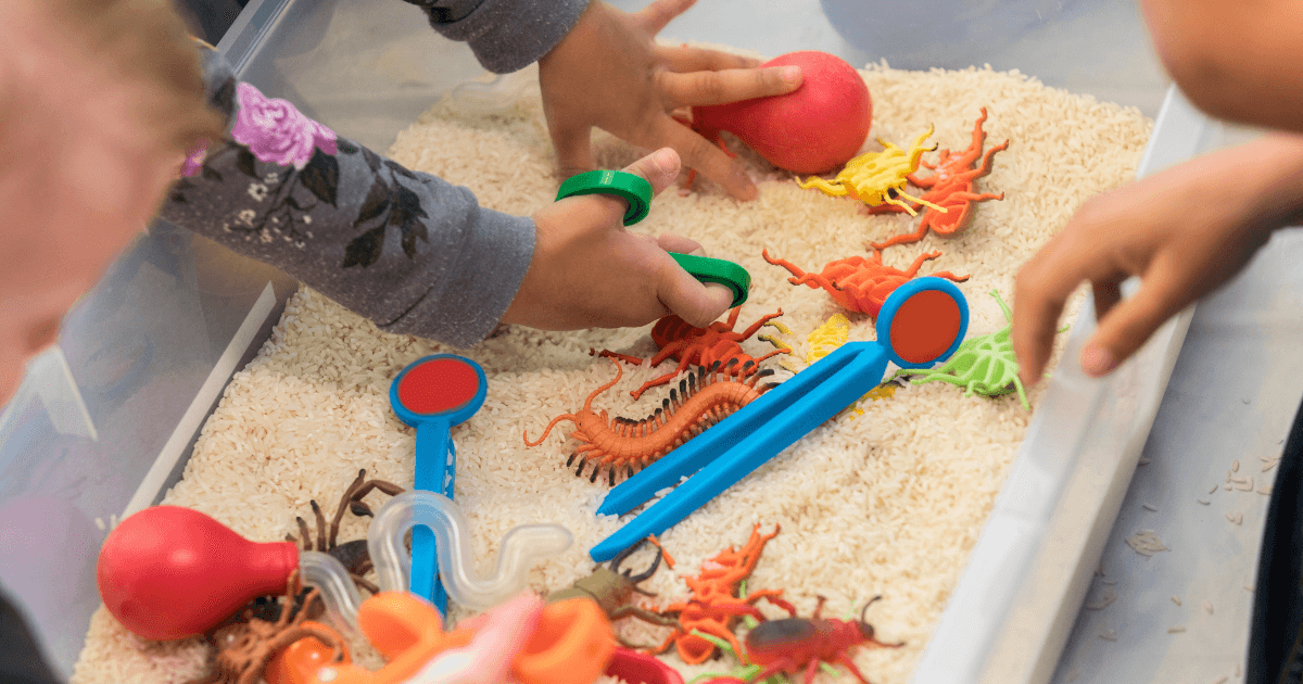Children’s hands playing with toy insects and colorful tweezers in a bin filled with dry rice, enjoying a seasonal sensory bins activity that fosters both fun and learning.