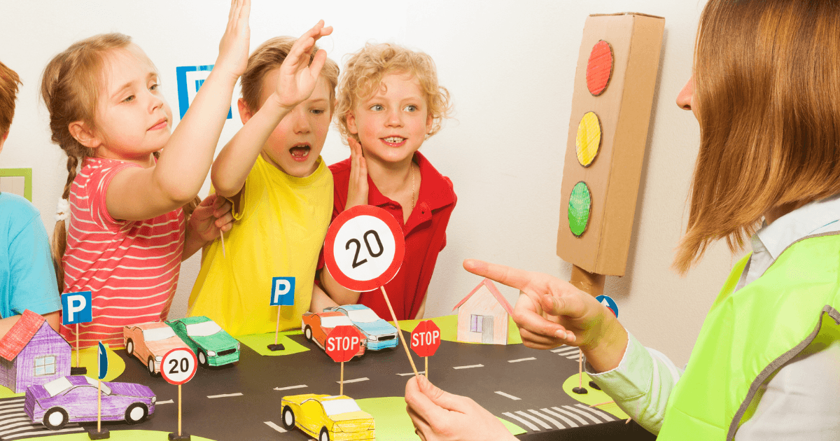Four young children enthusiastically raise their hands while learning about road safety and self-regulation in preschoolers from an adult. The scene includes toy cars, stop signs, parking signs, and a model traffic light on a table.