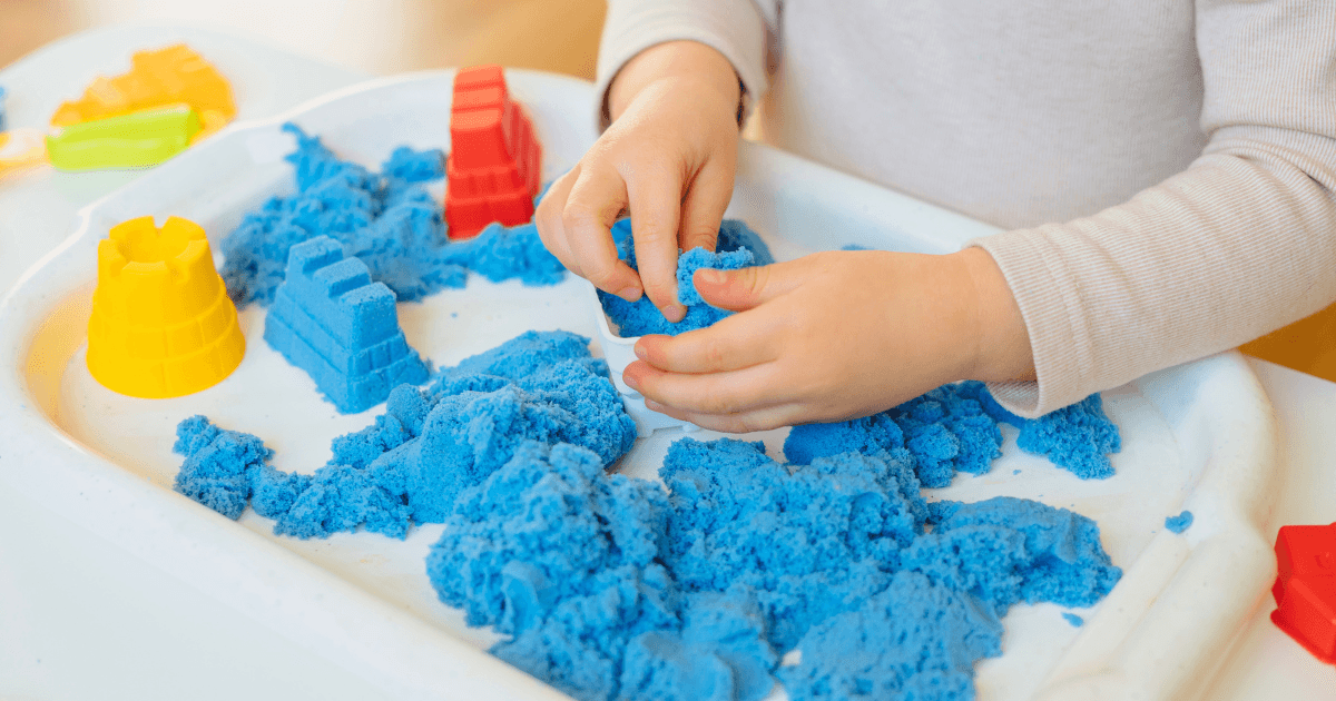 A child’s hands play with bright blue kinetic sand on a white tray, exploring sensory activities for preschoolers by molding it and using colorful plastic molds, including yellow and red shapes, to create different forms.
