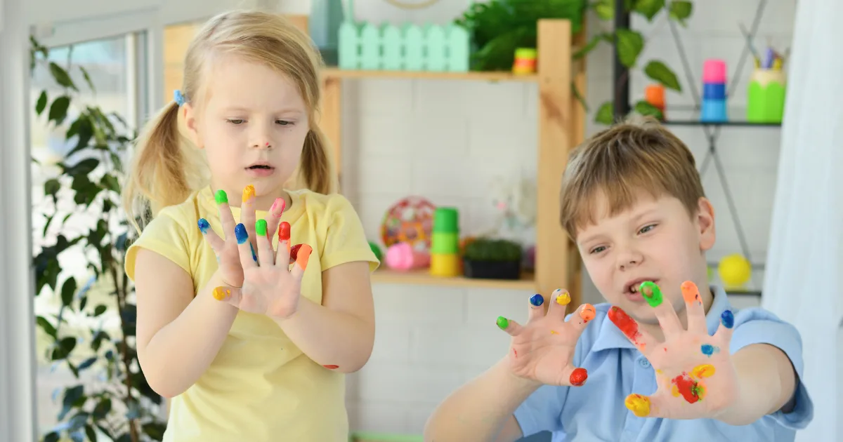 Two young children indoors with colorful paint on their fingers explore sensory processing as they hold up their hands and observe the paint. Art supplies and toys are visible in the background.