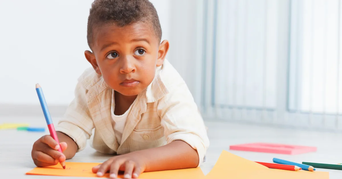 A young child lies on the floor, drawing on orange paper with a blue pencil. Colorful sheets of paper are scattered around as they look up thoughtfully, perhaps reflecting on questions like, "Should my child repeat preschool?.