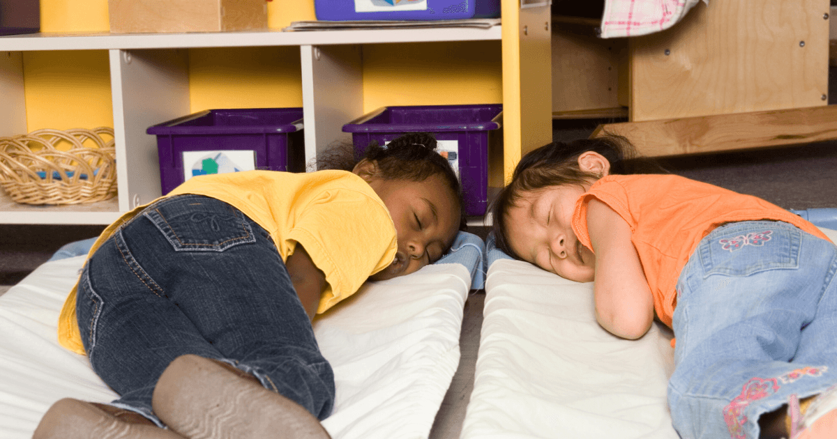 Two young children are lying on mats, napping side by side in a classroom setting, showing how sleep and learning go hand in hand, with shelves and bins in the background.