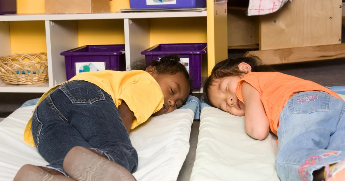 Two young children are lying on mats, napping side by side in a classroom setting, showing how sleep and learning go hand in hand, with shelves and bins in the background.