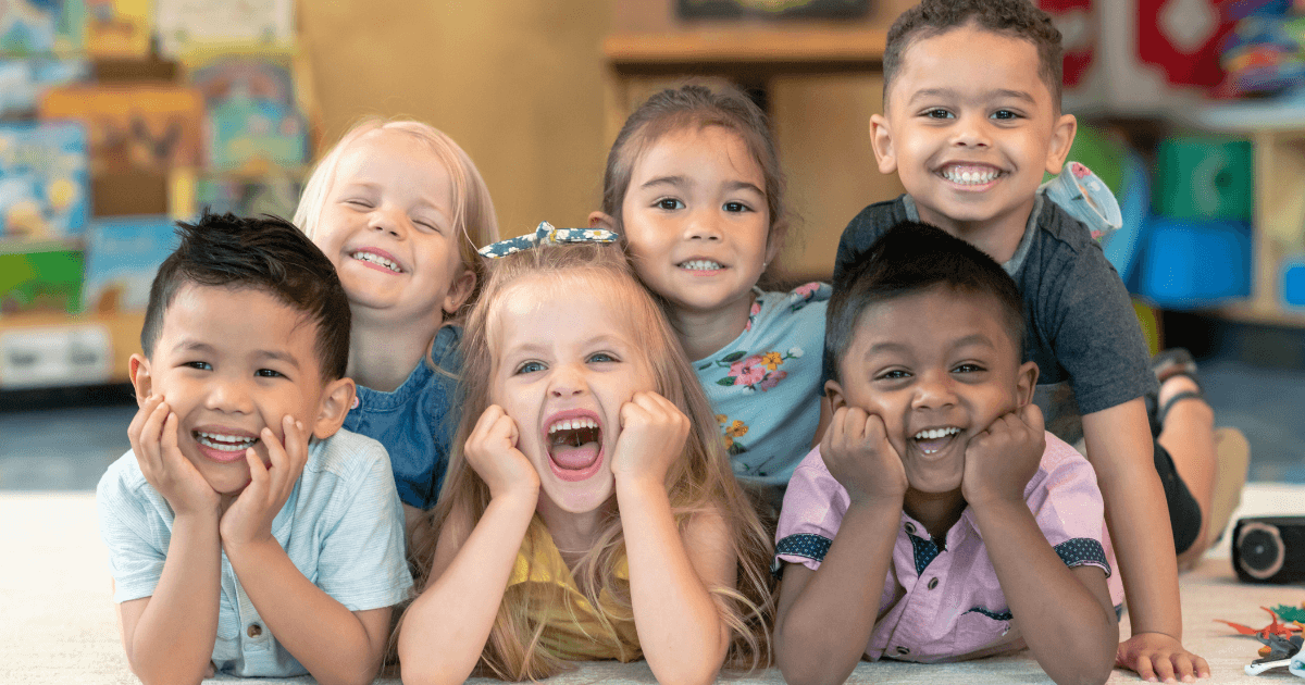 Six young children sit closely together on the classroom floor, smiling and laughing cheerfully at the camera—a joyful moment that highlights their social development. Books and colorful toys are visible in the background.