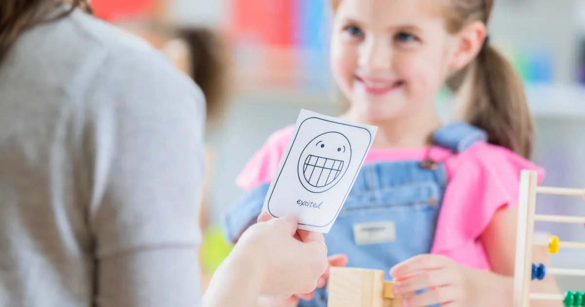 An adult holds a card with a drawing of a smiling face labeled excited while a young girl in pink and blue overalls smiles in the background, suggesting a social emotional learning or therapy activity.