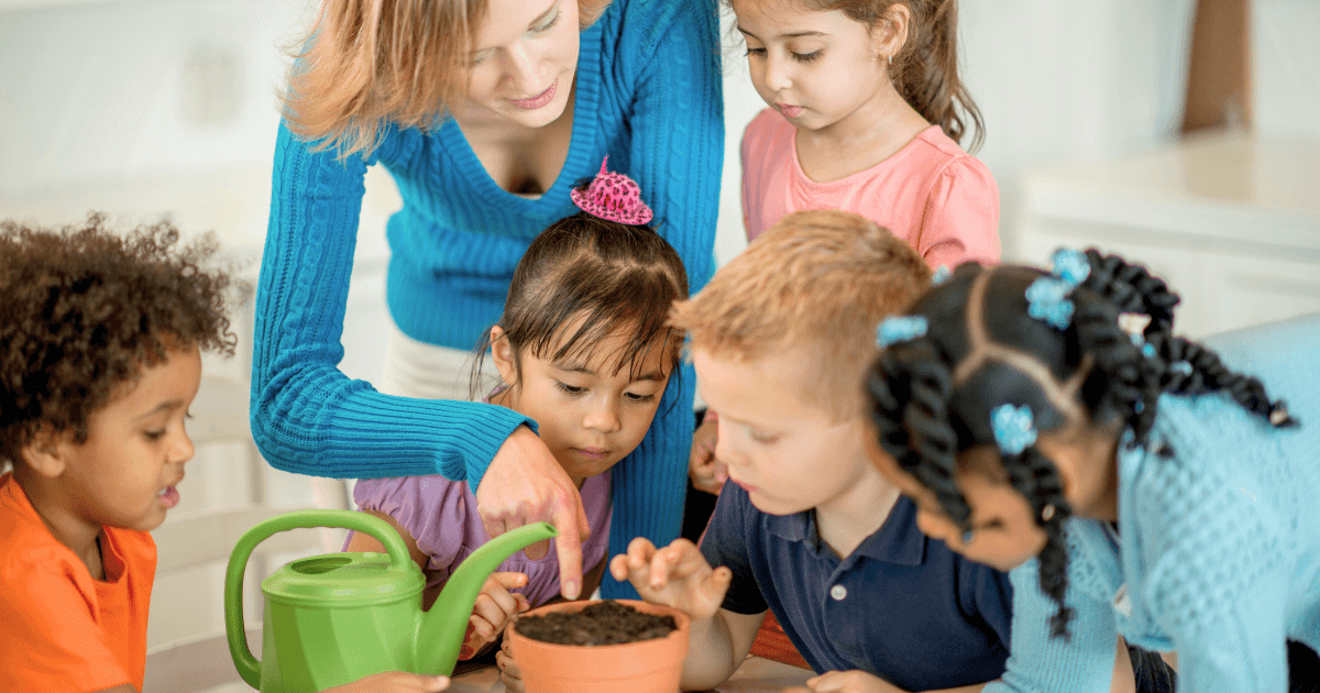 A woman and five young children gather around a table, closely examining a small potted plant as part of their spring planting projects. One child pours water from a green watering can while others touch the soil, engaging in gardening together.