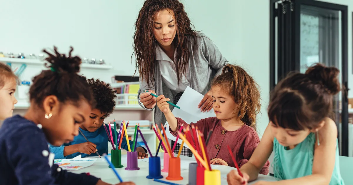 A teacher, key to staff retention, assists a group of young children engaged in drawing and coloring with colored pencils at a classroom table filled with various art supplies.