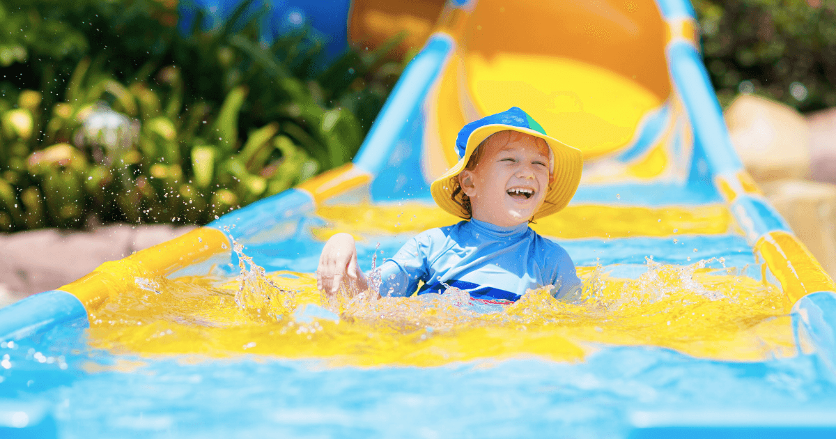 A young child wearing a yellow hat and blue swimsuit enjoys water play, splashing joyfully at the bottom of a bright blue and yellow water slide, with greenery in the background.