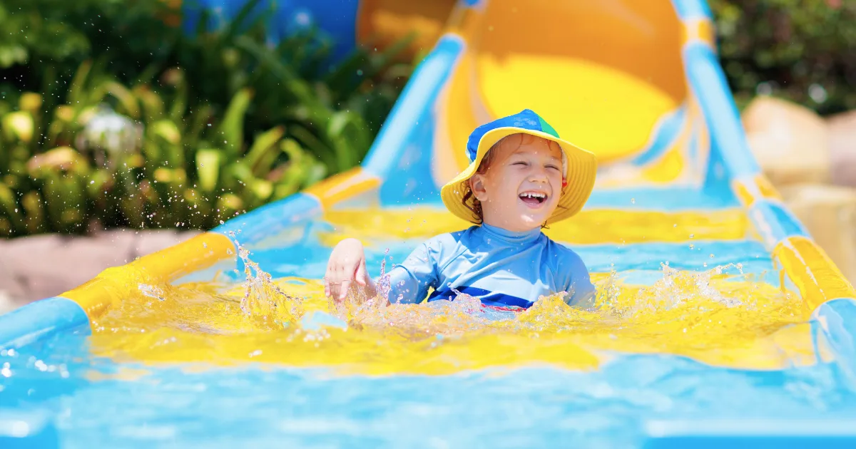 A young child wearing a yellow hat and blue swimsuit enjoys water play, splashing joyfully at the bottom of a bright blue and yellow water slide, with greenery in the background.