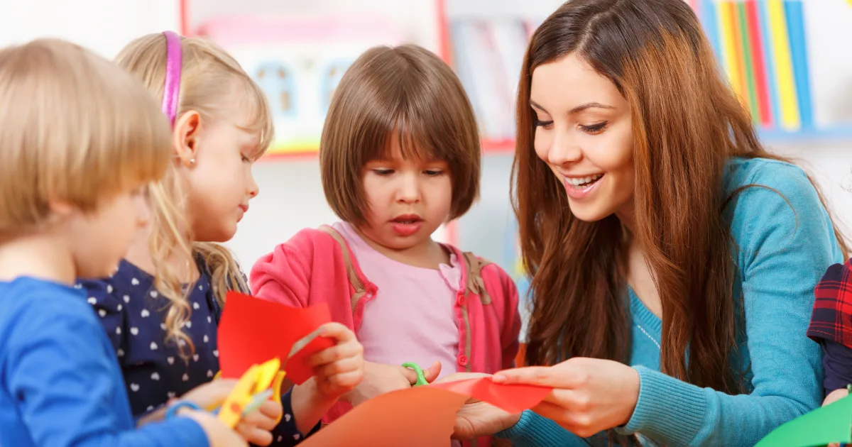 A young woman helps three children, including an anxious child, cut and fold colorful paper at a table. They smile and enjoy a creative activity together in a bright classroom setting.