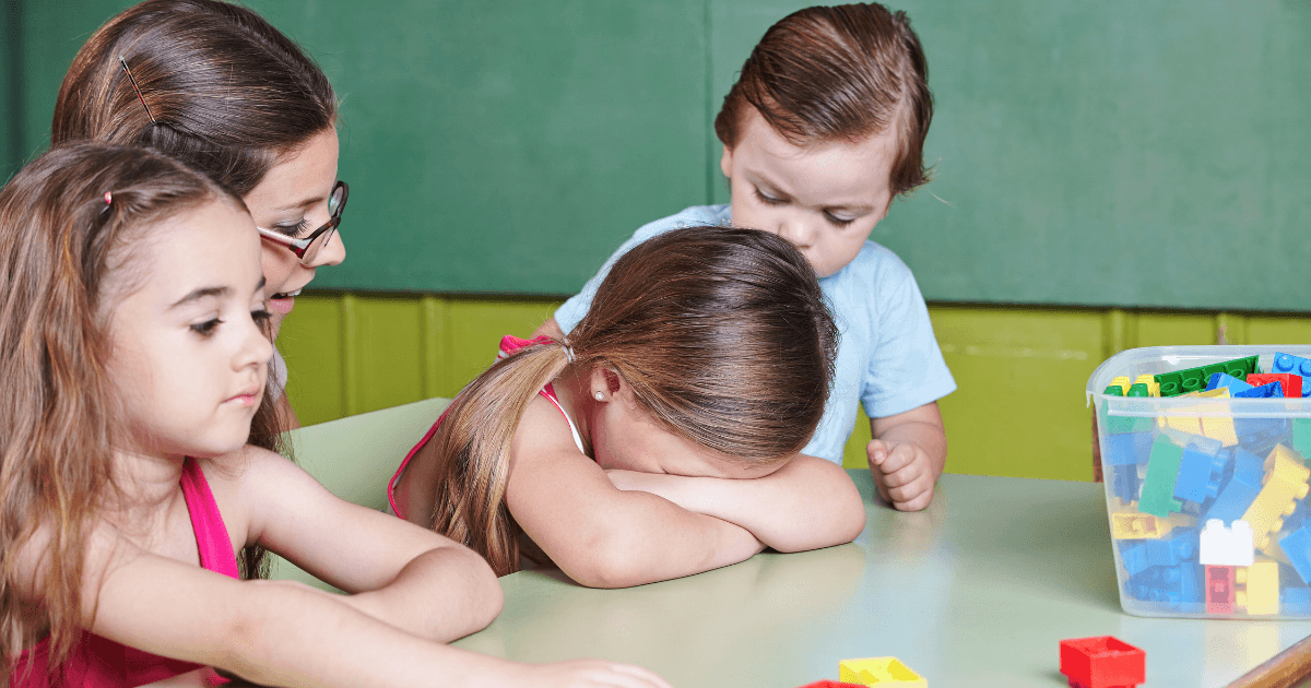 Four young children sit at a table with colorful plastic blocks. One girl has her head down on her arms, while another child looks concerned and two more look on, supporting children as they play or watch nearby.