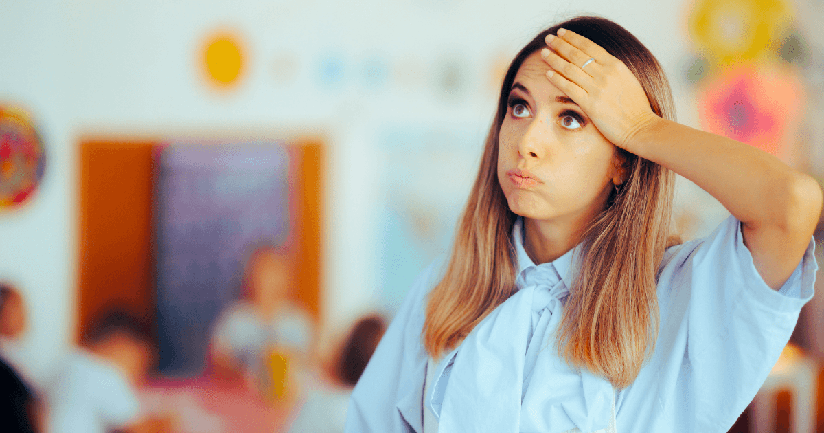 A young woman with long hair stands in a classroom, wearing a light blue shirt, holding her hand to her forehead and looking up with a frustrated or overwhelmed expression—highlighting the importance of teacher self-care. Other people are blurred in the background.
