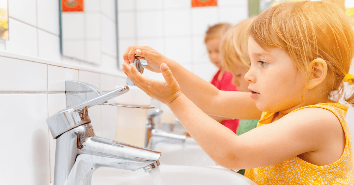 A young child in a yellow shirt practices hand washing at a sink, while two other children wash their hands in the background. The scene takes place in a bright, tiled bathroom.
