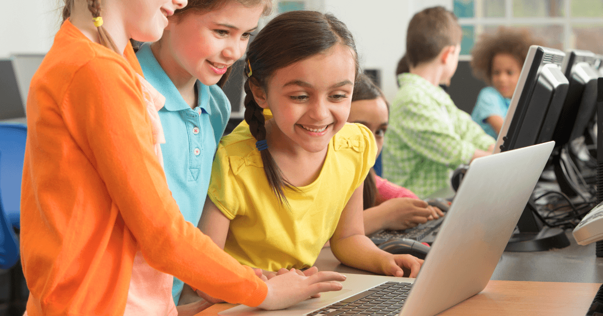 Three young girls smiling and working together on a laptop in a classroom, showcasing the use of technology in preschool, while other children engage with desktop computers in the background.