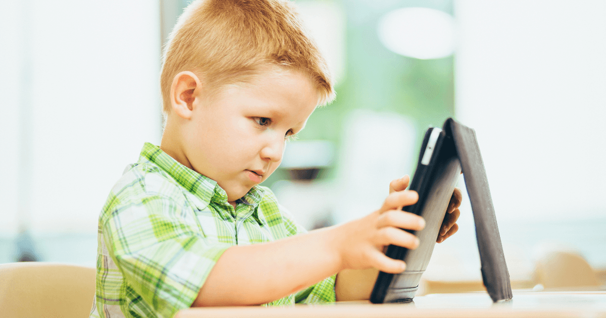 A young boy with blonde hair wearing a green plaid shirt sits at a table, focused on using a tablet with a black case—the digital preschooler in a bright, softly blurred background.