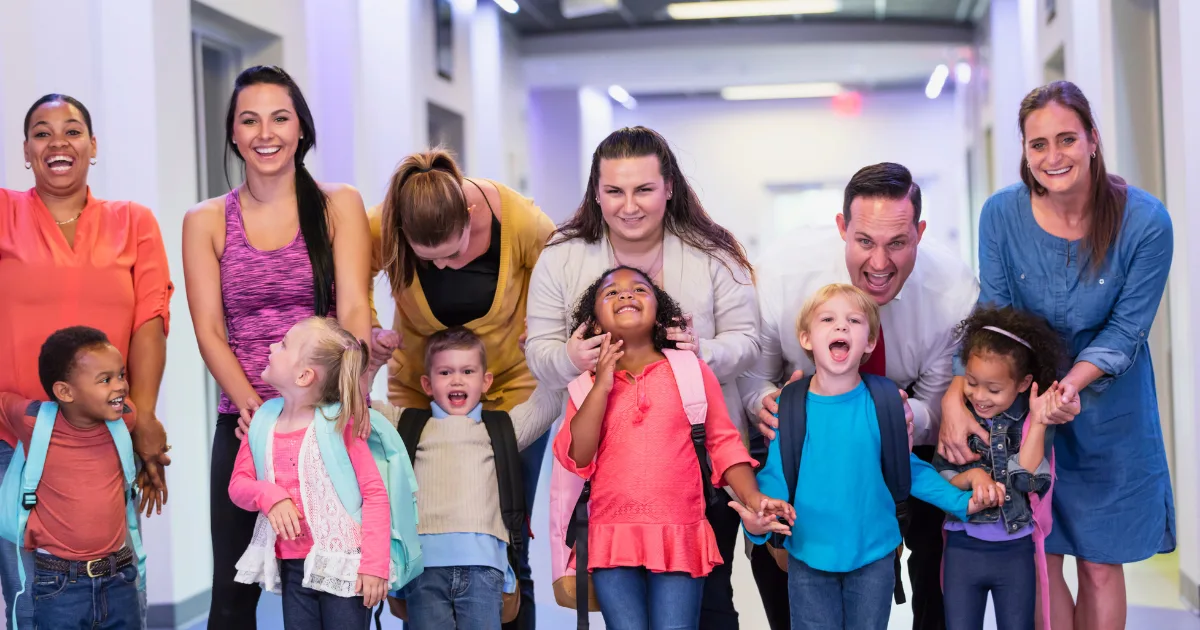 A group of smiling adults and young children, some wearing backpacks, stand together in a brightly lit hallway, laughing and holding hands during the first 30 days of preschool.