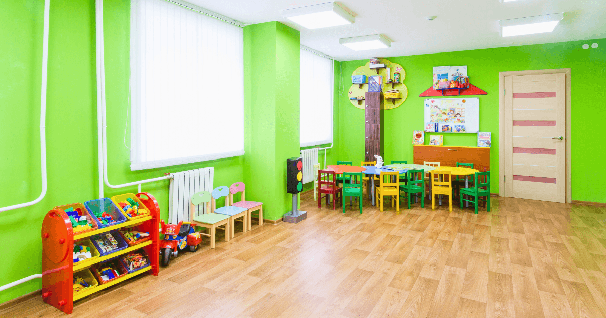 A brightly lit preschool environment with green walls, colorful children's chairs and tables, shelves with toys and books, and a wooden floor. Large windows let in natural light. The room looks tidy and inviting for kids.