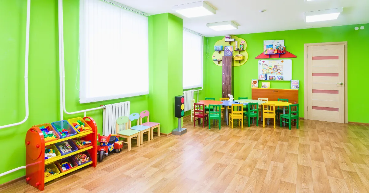 A brightly lit preschool environment with green walls, colorful children's chairs and tables, shelves with toys and books, and a wooden floor. Large windows let in natural light. The room looks tidy and inviting for kids.