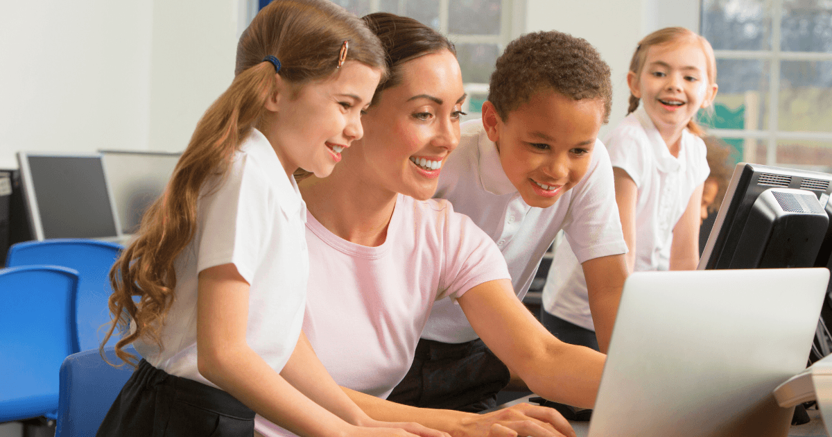 A teacher and three young students, all smiling, gather around a computer in a classroom with other computers and large windows—exploring time-saving documentation tools together.