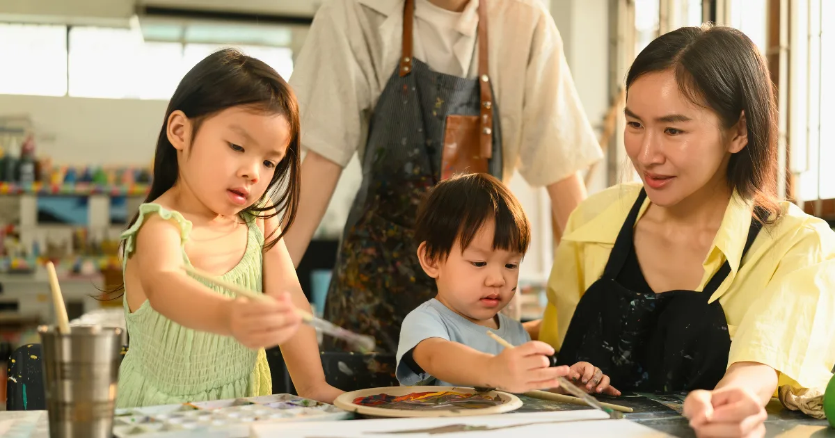 A woman and two young children sit at a table painting together in a bright, creative studio, capturing the engaging atmosphere of a traditional academic preschool as an adult stands nearby watching their focused artwork.