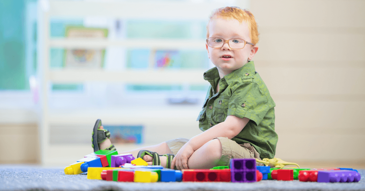 A toddler to preschool-aged boy with glasses and red hair sits on the floor, surrounded by colorful plastic building blocks, in a bright, indoor play area.