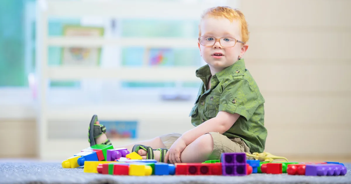 A toddler to preschool-aged boy with glasses and red hair sits on the floor, surrounded by colorful plastic building blocks, in a bright, indoor play area.