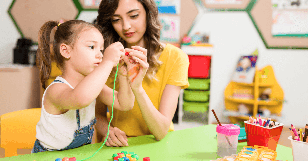 A young girl and a woman sit at a table in a colorful classroom, stringing large plastic beads together as part of transition techniques. Art supplies, cups, and containers fill the table, with bright shelves in the background.