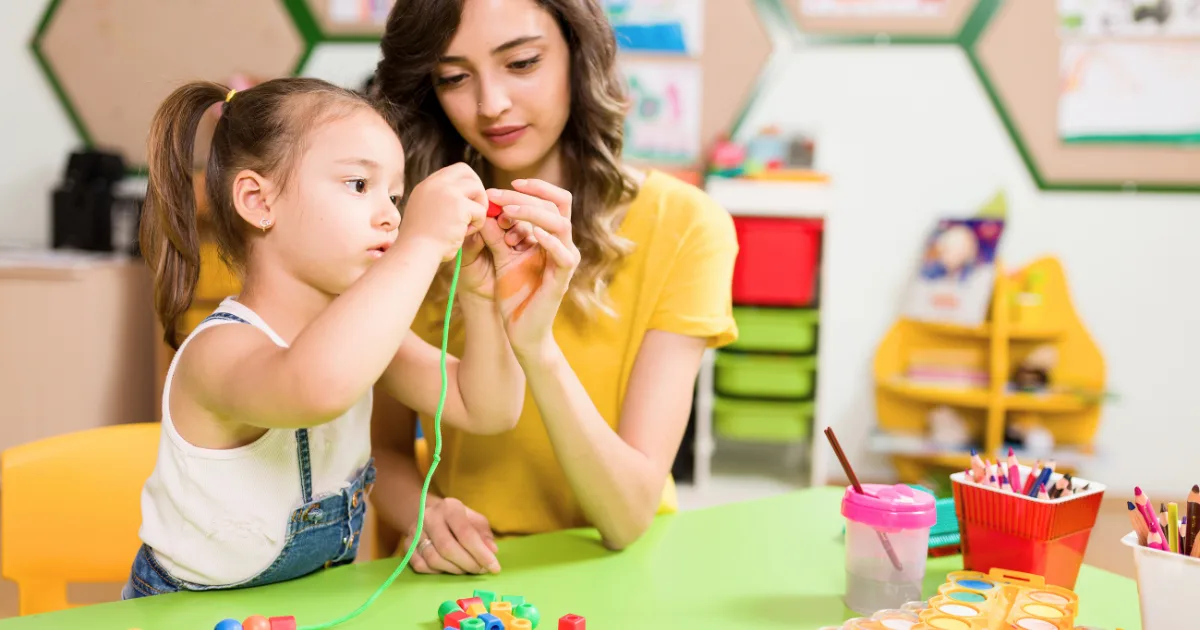 A young girl and a woman sit at a table in a colorful classroom, stringing large plastic beads together as part of transition techniques. Art supplies, cups, and containers fill the table, with bright shelves in the background.